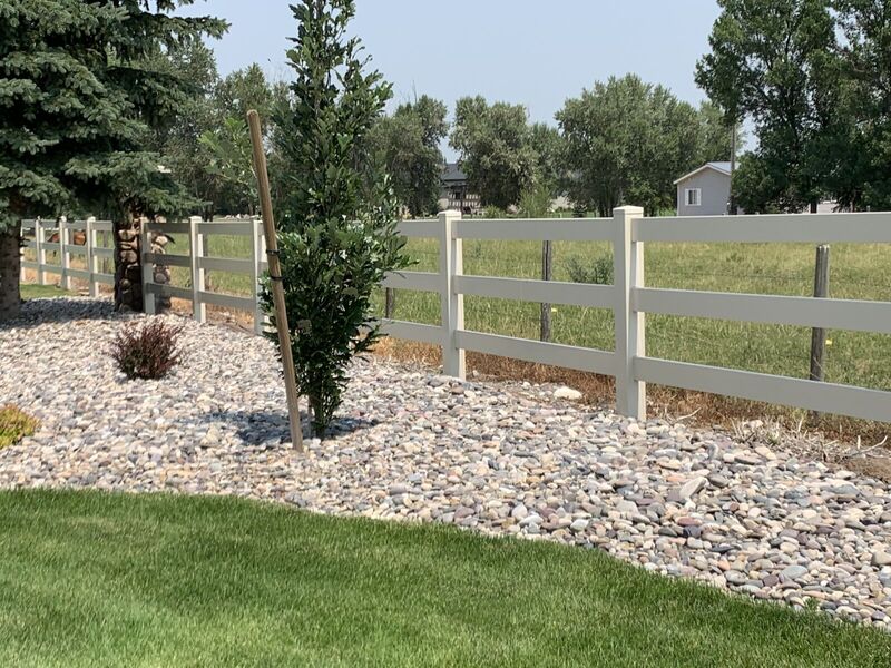 Tan vinyl three-rail ranch fence beside a river-rock landscape bed